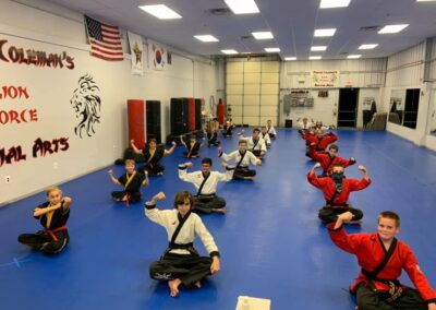 A group of martial arts students, both kids and adults, are sitting in rows on a blue mat in a martial arts dojo. They are all dressed in black and red uniforms, smiling, and striking a flexing pose with one arm. The dojo has various flags and martial arts decorations on the walls.