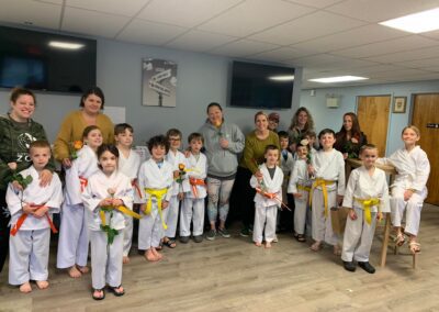 A group photo of children and adults in a martial arts studio. The children are dressed in white karate uniforms with various colored belts, while the adults stand behind them, some holding flowers. Everyone is smiling and looking at the camera.