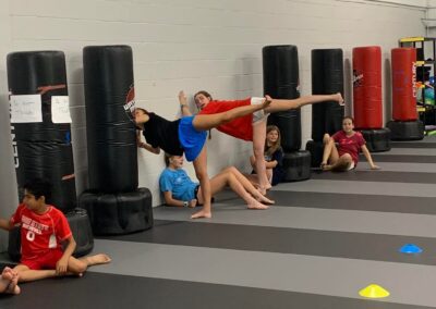 Children practice martial arts indoors, with some kicking standing punching bags while others sit on the padded floor watching. Colored cones and various sectioned areas are visible in the background, highlighting an organized training environment.