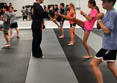 A martial arts class practicing kicks in a gym. An instructor in a black uniform supervises students as they perform front kicks. Students wear casual athletic clothing and stand in lines on mats, with mirrors on the walls reflecting the training.