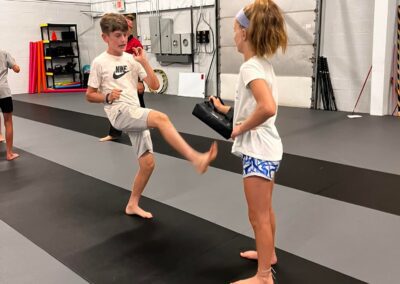 Two children are practicing martial arts in a gym. The boy on the left is delivering a front kick to a shield held by the girl on the right. Both are dressed in athletic wear and stand on a black and gray mat with gym equipment in the background.