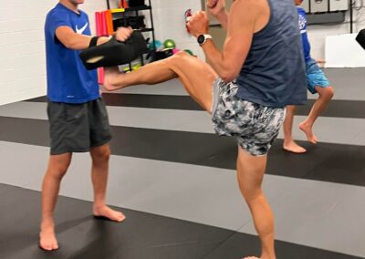 A woman wearing a tank top and shorts is performing a high kick into a pad held by a young boy in a gym. Another boy is observing in the background. They are all barefoot on a black-and-gray mat. The gym walls are white with framed photos and equipment visible.