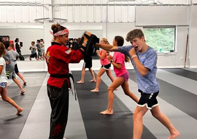 A group of teenagers practice martial arts in a studio with padded floors. An instructor in a red uniform holds pads for a boy in a blue shirt and black shorts to punch. Other students in the background work with partners. The studio has large windows and mirrors.