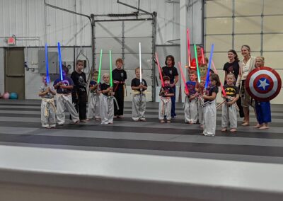 A group of children in martial arts uniforms hold colorful light sabers, standing on a mat in a gymnasium with a few adults. One child has a Captain America shield, and another adult is holding a light saber. They appear to be posing for a photo.