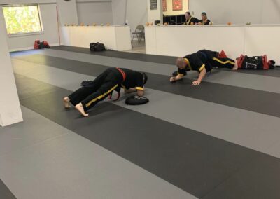 Two individuals dressed in martial arts uniforms are practicing on a gray and black matted floor inside a training facility. One person is on their hands and knees while the other is lying down, both focusing on an exercise. Mirrors line one wall of the room.