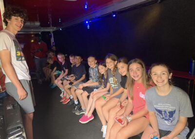 A group of teenagers sitting in a dark room lit with colorful lights, holding video game controllers. They are all sitting in a row on a bench, smiling and looking at the camera. One person is standing to the left, also smiling.