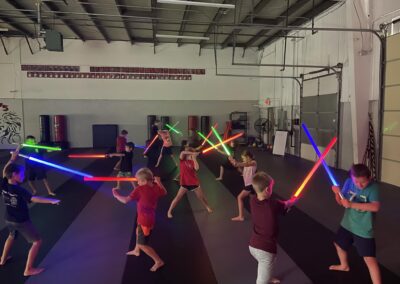 A group of children in a large indoor space engage in a mock lightsaber battle, each wielding brightly colored, glowing lightsabers. The room has high ceilings, and the children are spread out in pairs or small groups, all barefoot and actively participating.