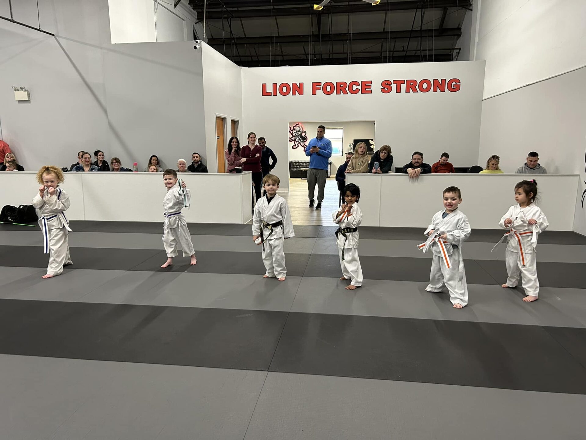 a group of children wearing white karate uniforms, standing in a line on a mat in a martial arts studio