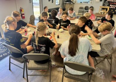 A group of children sits around a large table engaged in crafting activities. Various art supplies are spread across the table. The room has large windows, and motivational martial arts posters are visible on the walls.