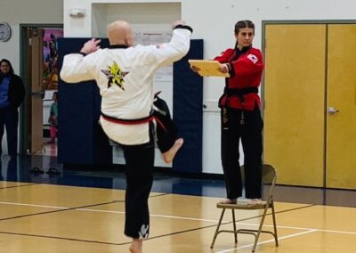 A person dressed in a martial arts uniform is in mid-air, performing a kick towards a board held by another person standing on a chair. The board holder is also wearing a martial arts uniform. The scene takes place in an indoor gymnasium.