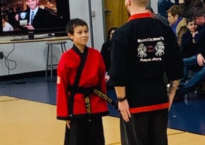 A young boy in a red martial arts uniform stands on a gymnasium floor, facing an adult instructor in a black martial arts uniform. A TV screen in the background shows a news broadcast. Spectators are seated along the wall, observing the interaction.