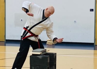 A martial artist in a white uniform with a red and black belt is captured mid-strike, attempting to break stacked concrete slabs on cinder blocks in a gymnasium. Concentration is evident on his face as he brings down his hand.