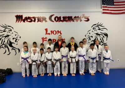 A group of children and two adults in martial arts uniforms stand in formation inside a dojo. The children wear various belt colors, and a U.S. flag is visible in the top right corner. The backdrop has "Master Coleman's Lion Force" with stylized lion designs.