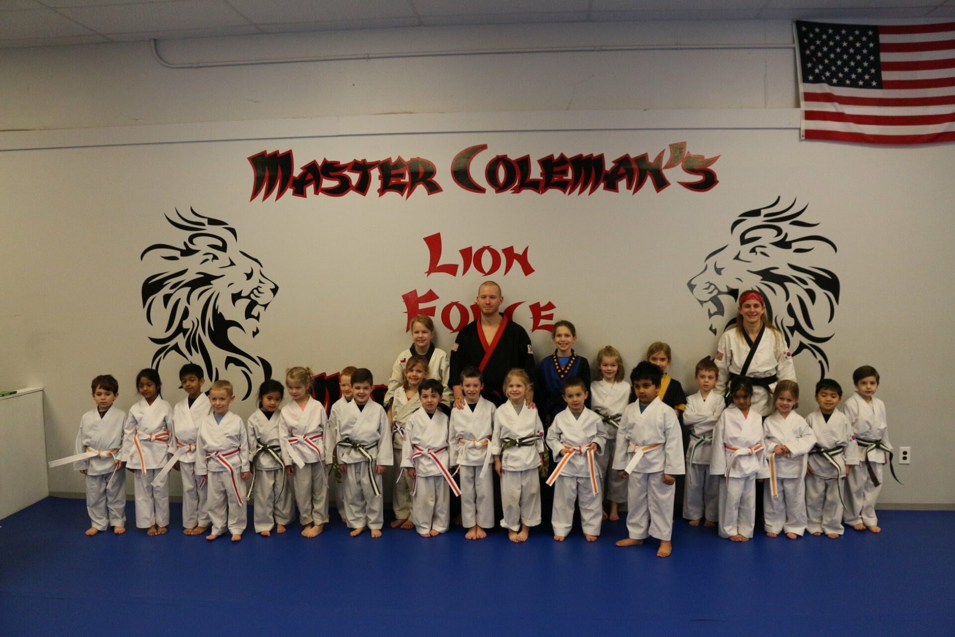 A group of children in martial arts uniforms stand in a row in front of a wall with the text "Master Coleman, Lion Force, SMA"